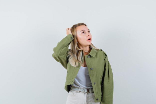 Young lady holding hand on head in jacket, pants and looking dreamy , front view.