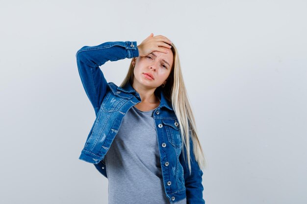 Young lady holding hand on head in t-shirt, denim jacket, skirt and looking tired