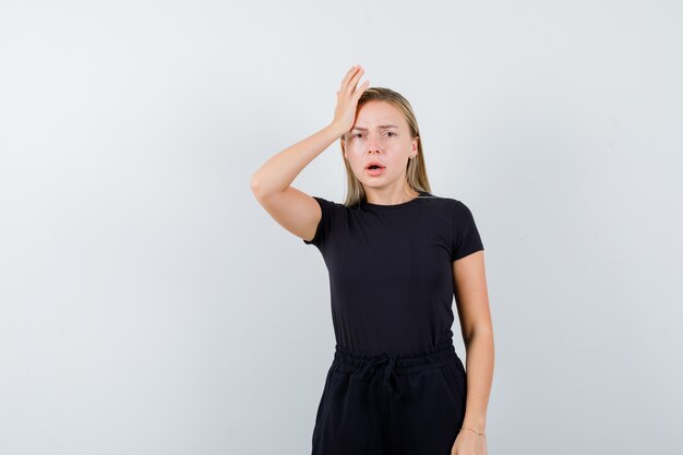 Young lady holding hand on head in t-shirt, pants and looking anxious. front view.