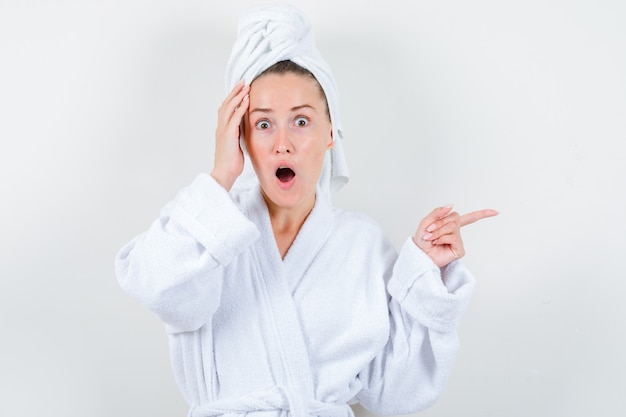 Young lady holding hand on head while pointing to the right side in white bathrobe, towel and looking surprised , front view.