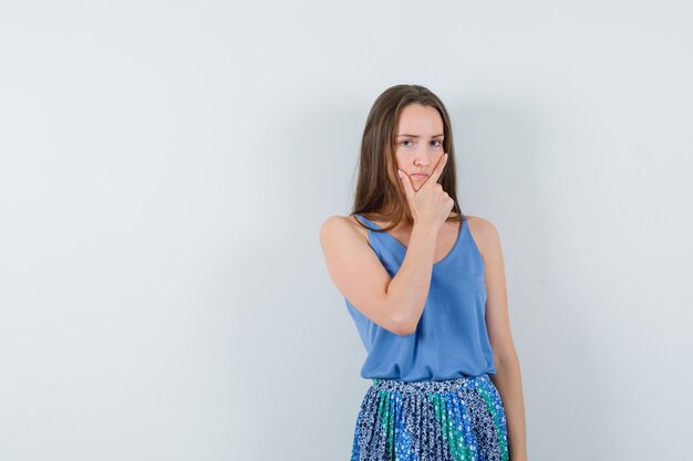 Young lady holding hand on her jaw while thinking in blue blouse,skirt and looking pensive , front view. space for text