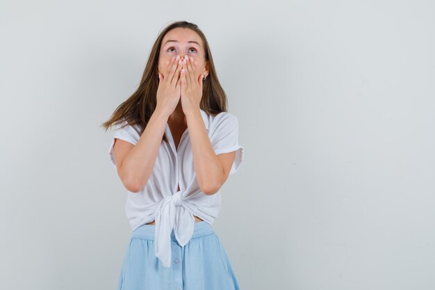 Young lady holding hand on her mouth while looking up in blouse, skirt and looking excited