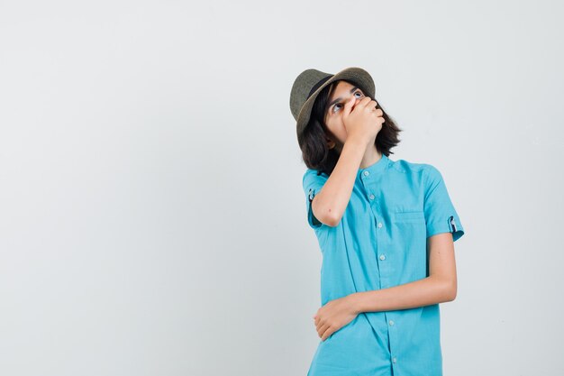 Young lady holding hand on her mouth while looking up in blue shirt, hat and looking scared.