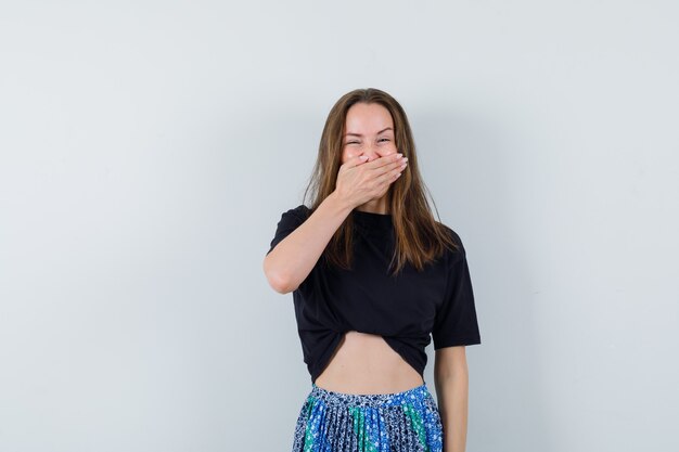 Young lady holding hand on mouth in blouse, skirt and looking happy