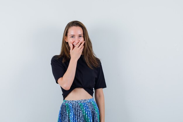 Young lady holding hand on mouth in blouse, skirt and looking merry