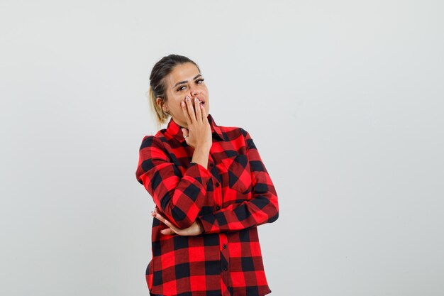 Young lady holding hand on mouth in checked shirt and looking surprised