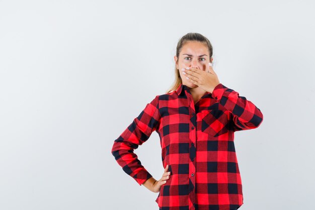 Young lady holding hand on mouth in checked shirt and looking surprised