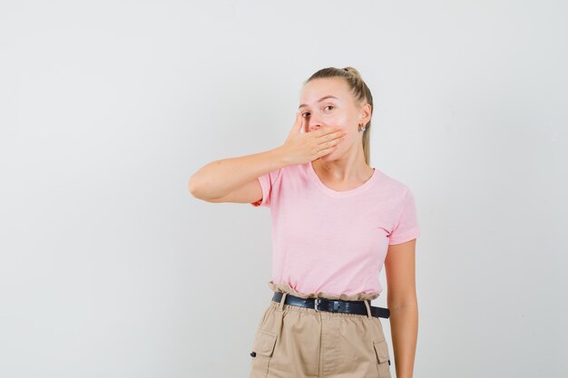 Young lady holding hand on mouth in t-shirt, pants and looking mysterious. front view.