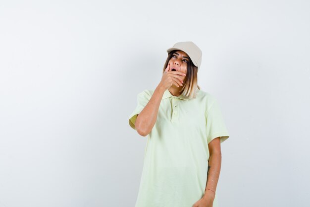 Young lady holding hand on mouth while looking up in t-shirt, cap and looking puzzled , front view.