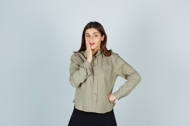 Young lady holding hand near mouth in shirt, skirt and looking pensive