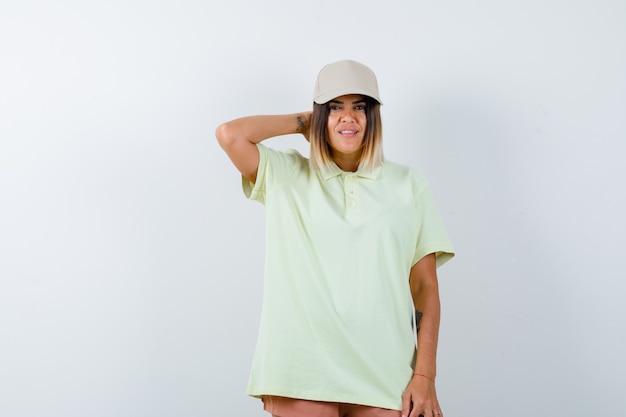 Young lady holding hand on neck while posing in t-shirt, cap, short and looking confident , front view.