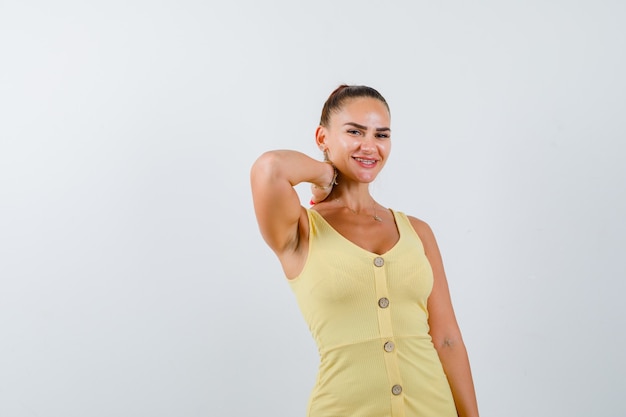 Foto gratuita giovane donna che tiene la mano dietro il collo in abito giallo e guardando allegro, vista frontale.