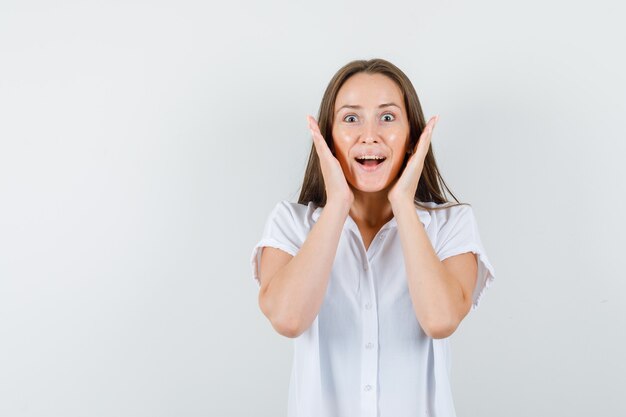 Young lady holding hands around her face in white blouse and looking glad.