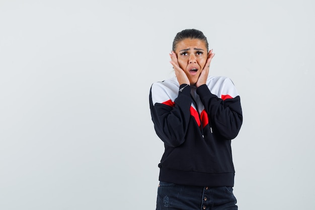 Young lady holding hands on cheek in colorful sweatshirt and looking scared , front view. 