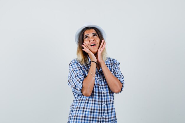 Young lady holding hands on cheeks in checked shirt hat and looking cute 