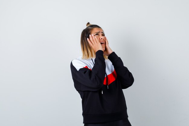 Young lady holding hands on cheeks in hoodie sweater and looking sorrowful.