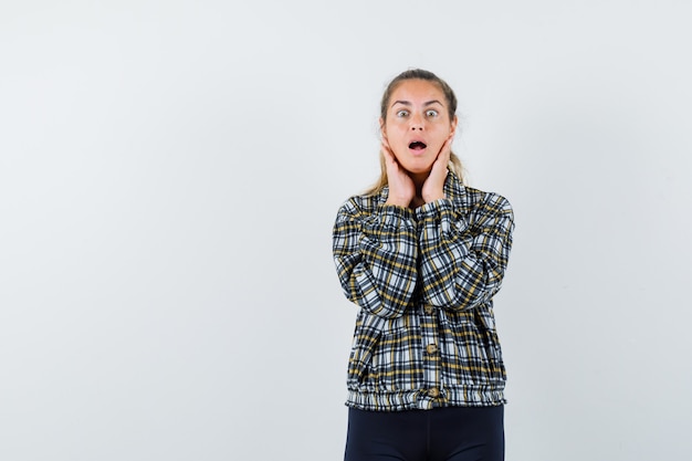 Young lady holding hands on cheeks in shirt, shorts and looking surprised , front view.