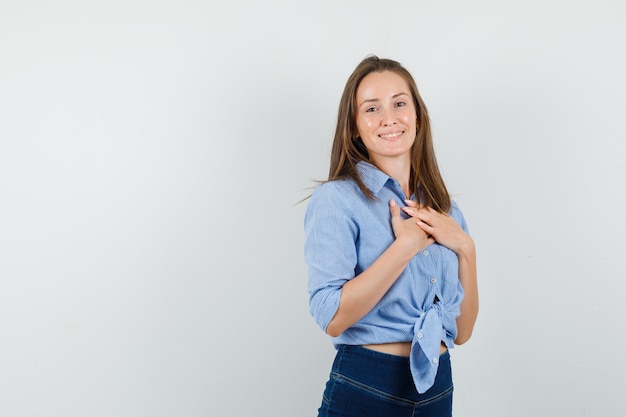 Young lady holding hands on chest in blue shirt, pants and looking cheerful