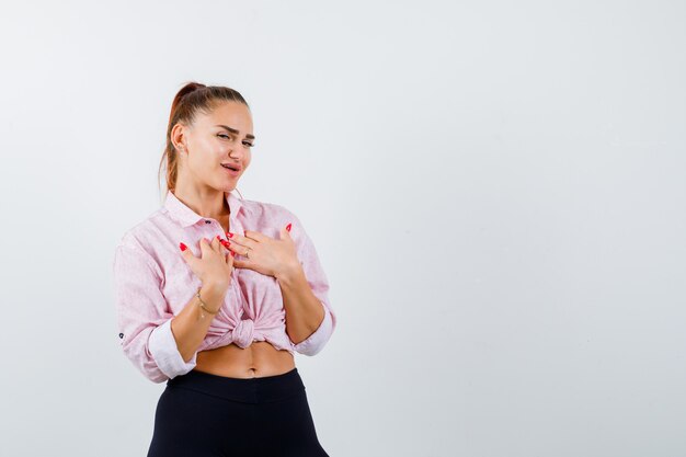 Young lady holding hands on chest in shirt, pants and looking grateful. front view.