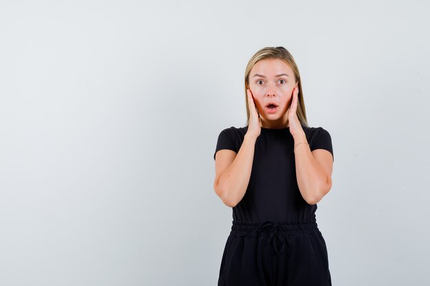 Young lady holding hands on ears in t-shirt, pants and looking surprised , front view.