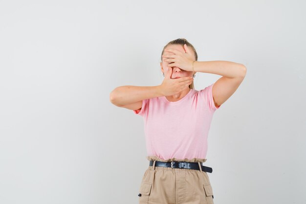 Young lady holding hands on eyes and mouth in t-shirts and pants and looking scared