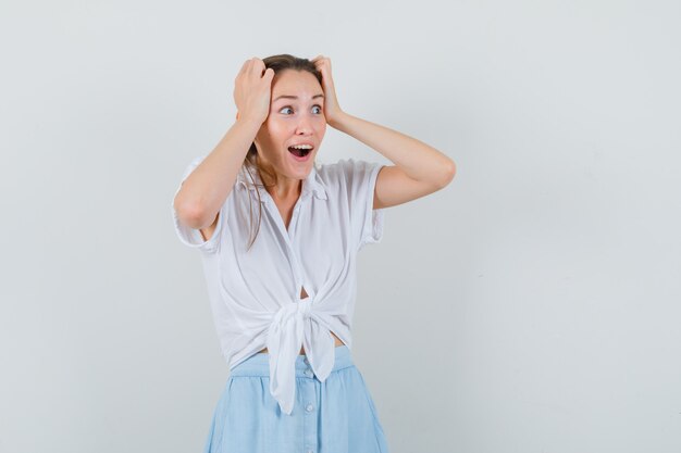 Young lady holding hands on head in blouse and skirt and looking lucky