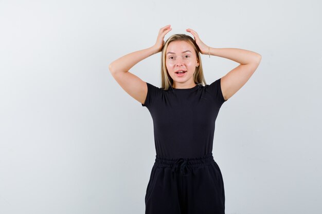 Young lady holding hands on head in t-shirt, pants and looking joyful , front view.