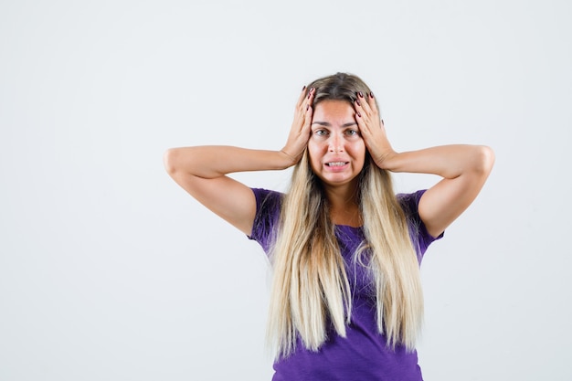 Young lady holding hands to head in violet t-shirt and looking wistful , front view.