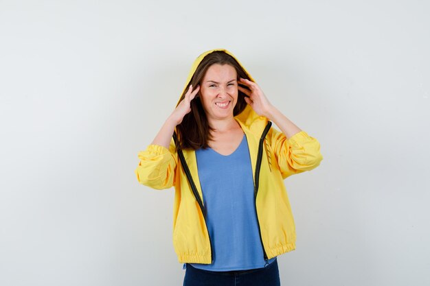 Young lady holding hands to head while frowning in t-shirt, jacket and looking confused