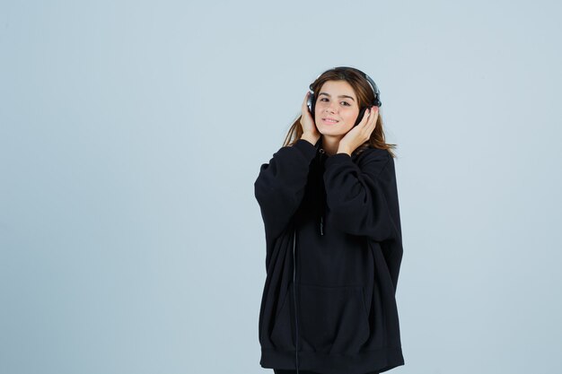 Young lady holding hands on headphones in oversized hoodie, pants and looking cheerful , front view.