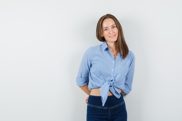 Young lady holding hands on her back in blue shirt, pants and looking optimistic