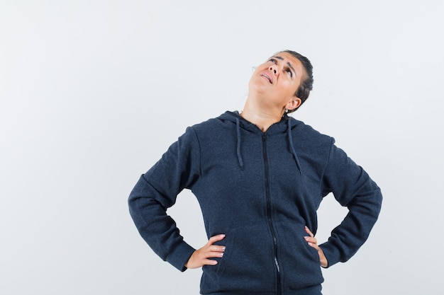 Young lady holding hands on her waist while looking up in jacket and looking focused. front view.