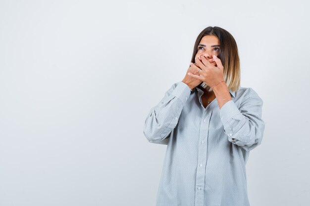 Young lady holding hands on mouth in oversize shirt and looking ashamed front view.