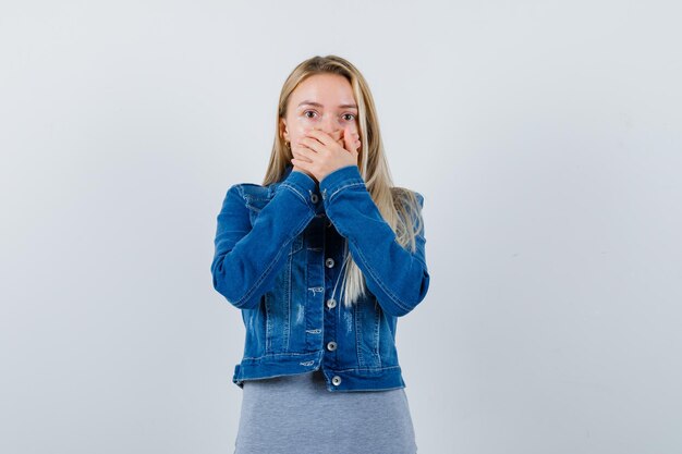 Young lady holding hands on mouth in t-shirt, denim jacket, skirt and looking amazed