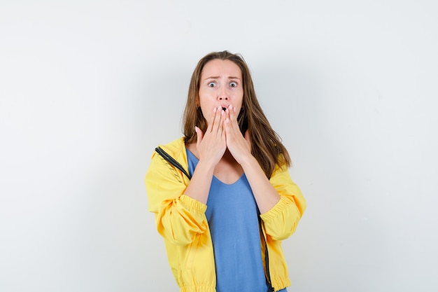 Young lady holding hands on mouth in t-shirt, jacket and looking shocked. front view.