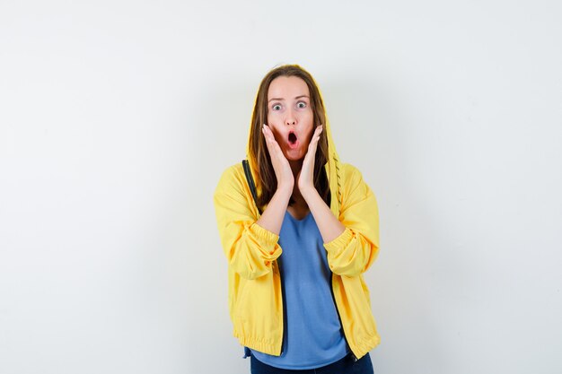 Young lady holding hands near open mouth in t-shirt, jacket and looking surprised. front view.