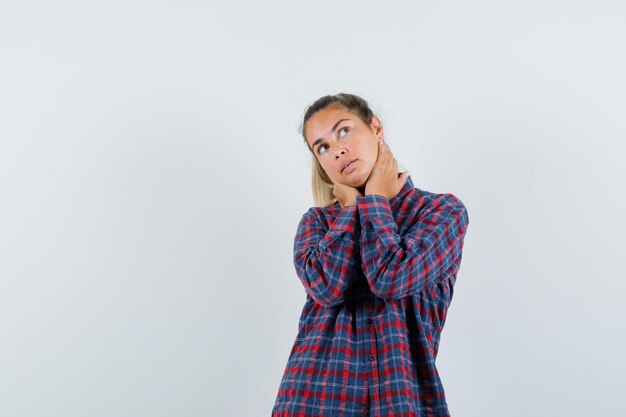 Young lady holding hands on neck in checked shirt and looking dreamy. front view.