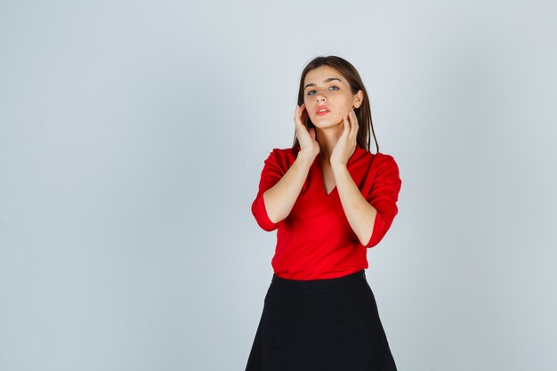 Young lady holding hands on neck in red blouse, black skirt and looking alluring