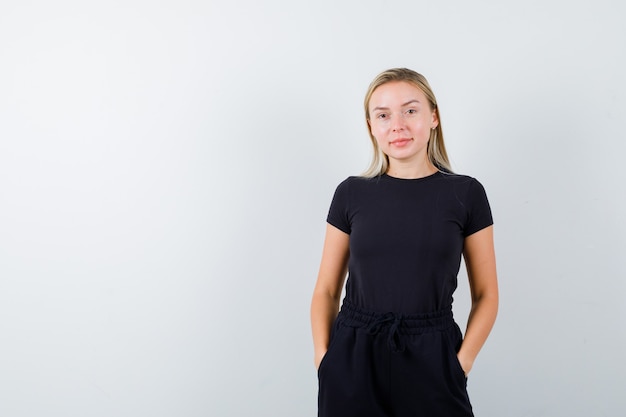 Young lady holding hands in pockets in t-shirt, pants and looking cheery. front view.