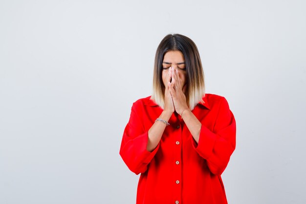 Young lady holding hands in praying gesture in red oversize shirt and looking hopeful. front view.