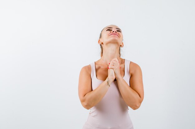 Young lady holding hands in praying gesture in singlet and looking hopeful. front view.