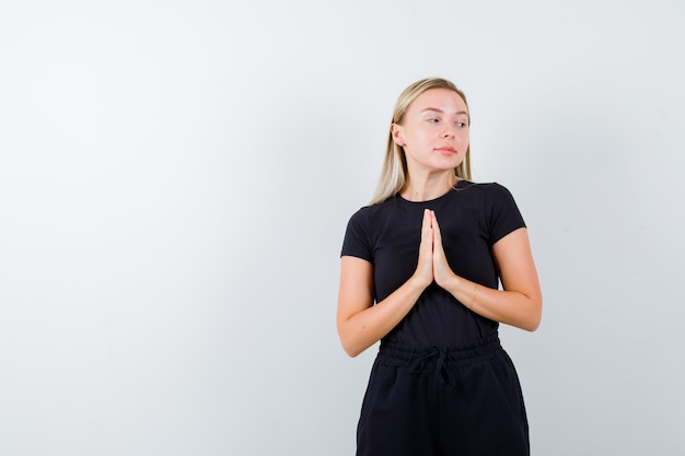 Young lady holding hands in praying gesture in t-shirt, pants and looking hopeful. front view