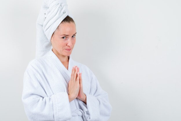Young lady holding hands in praying gesture in white bathrobe, towel and looking hopeful , front view.
