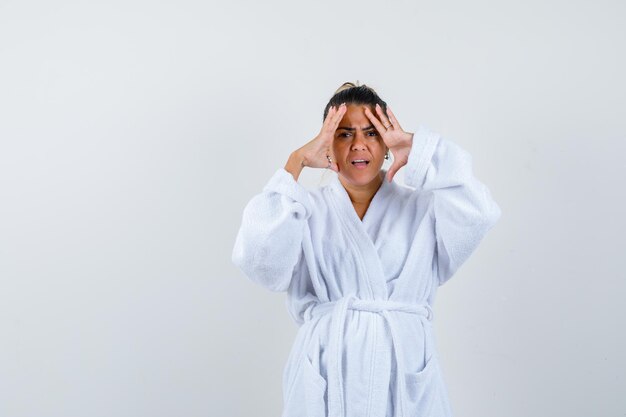 Young lady holding hands on temples in bathrobe and looking fatigued