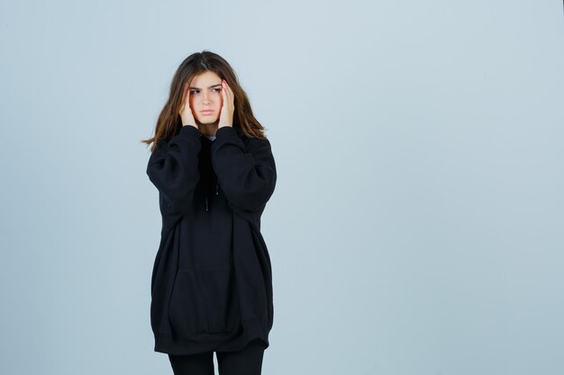 Young lady holding hands on temples in oversized hoodie, pants and looking fatigued. front view.