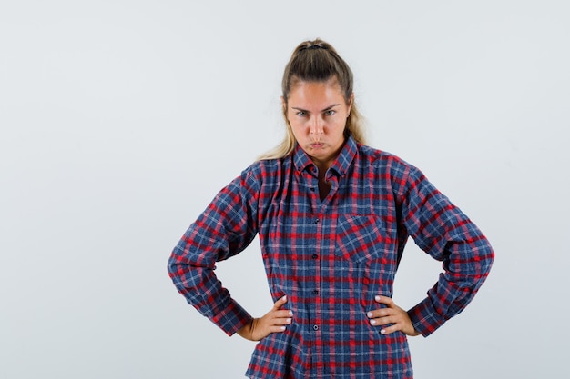 Young lady holding hands on waist in checked shirt and looking pensive. front view.