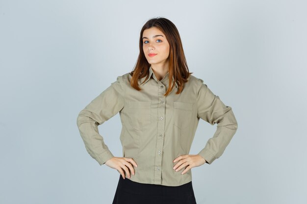 Young lady holding hands on waist in shirt, skirt and looking confident , front view.