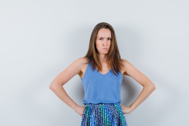 Young lady holding hands on waist in singlet, skirt and looking spiteful