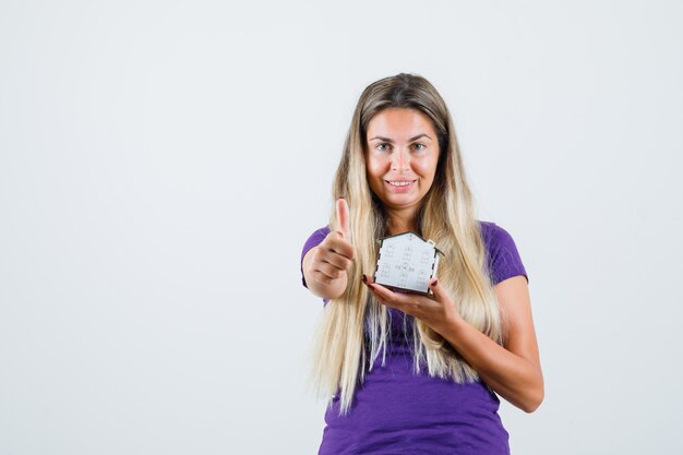 Young lady holding house model, showing thumb up in violet t-shirt and looking cheerful. front view.