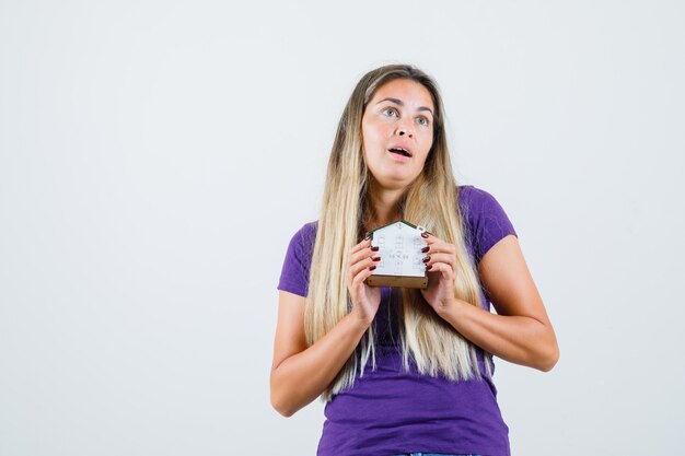 Young lady holding house model in violet t-shirt and looking surprised. front view.